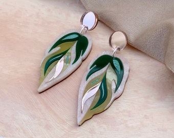 May include: A pair of leaf-shaped earrings. The earrings feature a layered design with shades of green, cream, and a touch of pink. The top of each earring has a small, round, silver-colored stud. The earrings are set against a light-colored background.