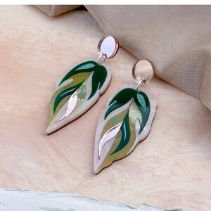 Könnte beinhalten: Ein Paar ohrförmige Ohrringe. Die Ohrringe haben ein geschichtetes Design mit Grüntönen, Creme und einem Hauch von Rosa. Die Oberseite jedes Ohrrings hat einen kleinen, runden, silberfarbenen Stecker. Die Ohrringe sind vor einem hellen Hintergrund platziert.