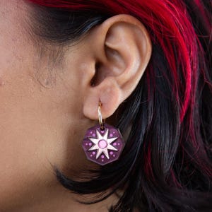 Könnte beinhalten: Nahaufnahme eines goldenen Creolen-Ohrrings mit einem lila, geometrischen Anhänger. Der Anhänger hat ein weißes Sternenmuster mit rosa Akzenten. Der Ohrring wird von einer Person mit roten und schwarzen Haaren getragen.