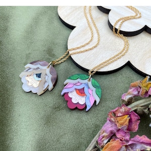 May include: Two gold chain necklaces with colorful flower-shaped pendants. One pendant is white, brown, and blue with an eye design. The other pendant is green, pink, red, and white with a flower design.