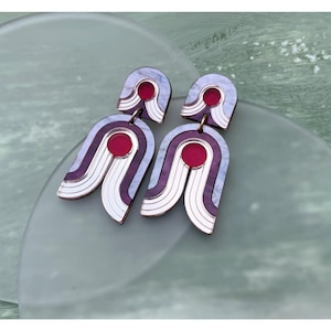 Könnte beinhalten: Ein Paar geometrische Ohrringe in Lila und Weiß mit roten Akzenten. Die Ohrringe sind aus Acryl und haben ein modernes, abstraktes Design.