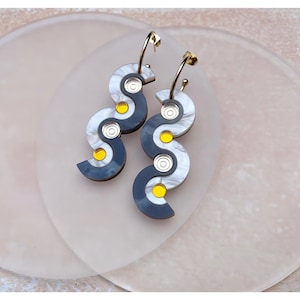 Könnte beinhalten: Ein Paar lange, baumelnde Ohrringe mit einem wellenförmigen, abstrakten Design. Die Ohrringe sind aus schwarzem und weißem Acryl mit goldenen Akzenten und gelben Kreisen.