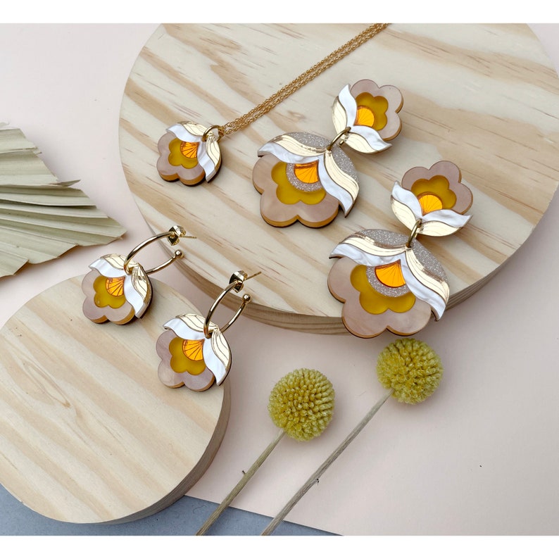 Pode incluir: Um conjunto de brincos florais dourados e brancos e um colar combinando. Os brincos s&atilde;o feitos de acr&iacute;lico em camadas e t&ecirc;m um centro amarelo. O colar &eacute; uma corrente simples com um &uacute;nico pingente floral.