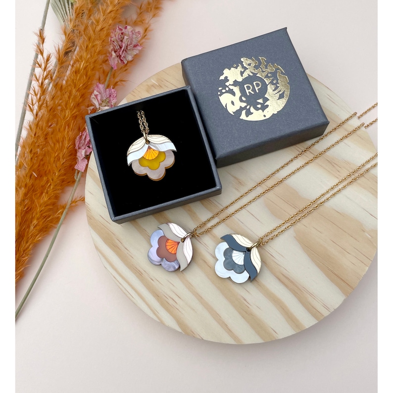 Pode incluir: Dois colares de ouro com pingentes em forma de flor. Um pingente &eacute; branco, laranja e marrom. O outro pingente &eacute; branco, cinza e preto. Os colares s&atilde;o exibidos em uma superf&iacute;cie de madeira com uma caixa cinza no fundo.