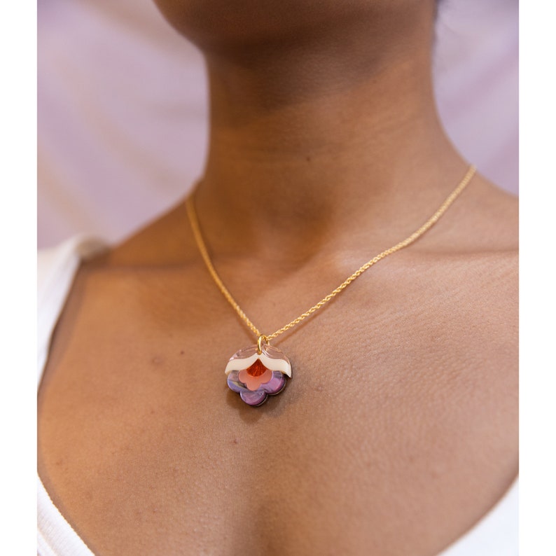 May include: A gold chain necklace with a layered flower pendant. The flower is made of translucent plastic and has shades of pink, orange, and purple.