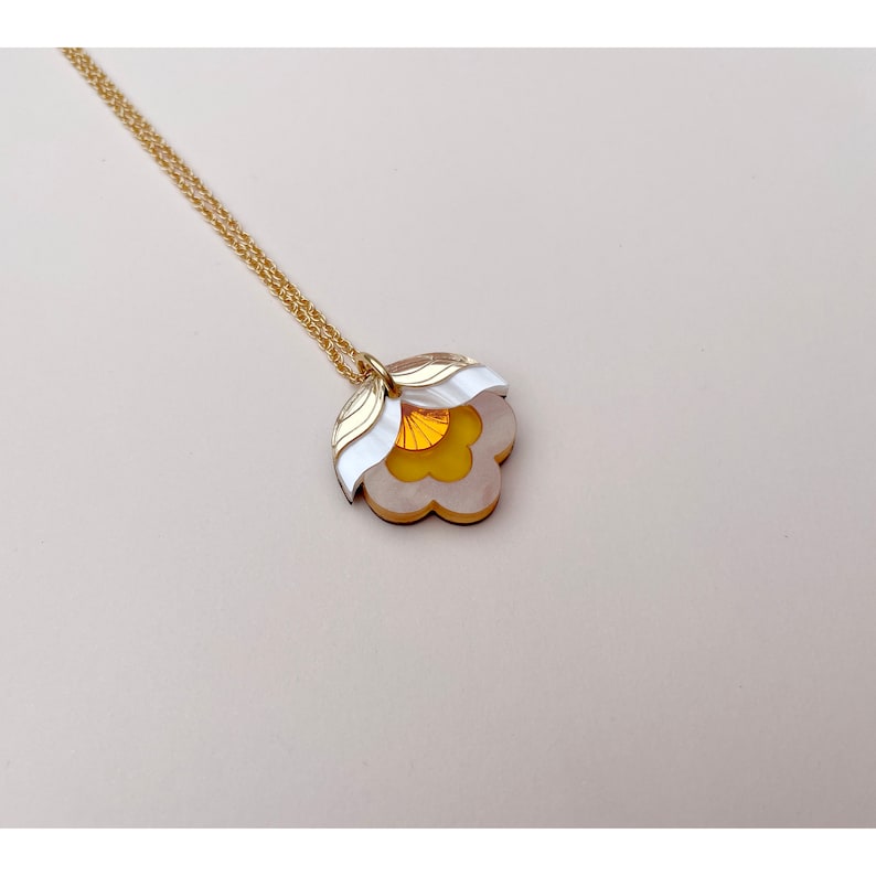 Pode incluir: Um colar de corrente dourada com um pingente de flor branca e amarela. A flor tem um design em camadas com um centro amarelo e p&eacute;talas brancas.