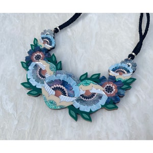 Könnte beinhalten: Eine auffällige Blumenkette mit stilisierten blau-cremefarbenen Blumen und grünen Blättern. Die Halskette hat eine schwarze Kordel. Die Blumen haben Glitzerakzente und sind vor einem weißen Hintergrund platziert.