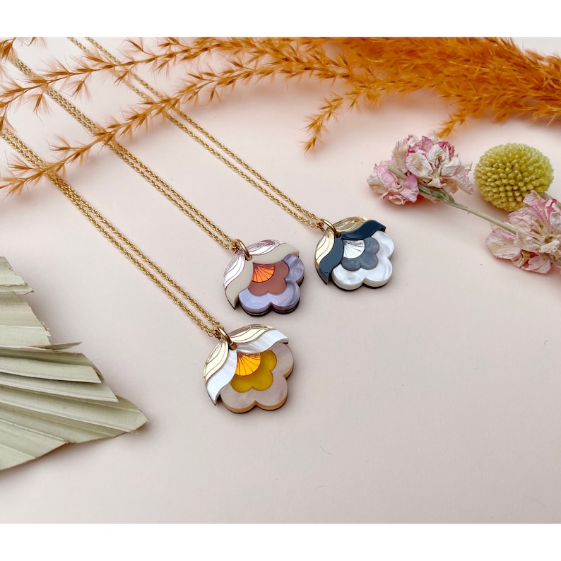 Pode incluir: Tr&ecirc;s colares de corrente dourada com pingentes de flores sobrepostas. Os pingentes s&atilde;o feitos de acr&iacute;lico de diferentes cores e t&ecirc;m um design geom&eacute;trico. Os colares s&atilde;o exibidos em uma superf&iacute;cie rosa claro.