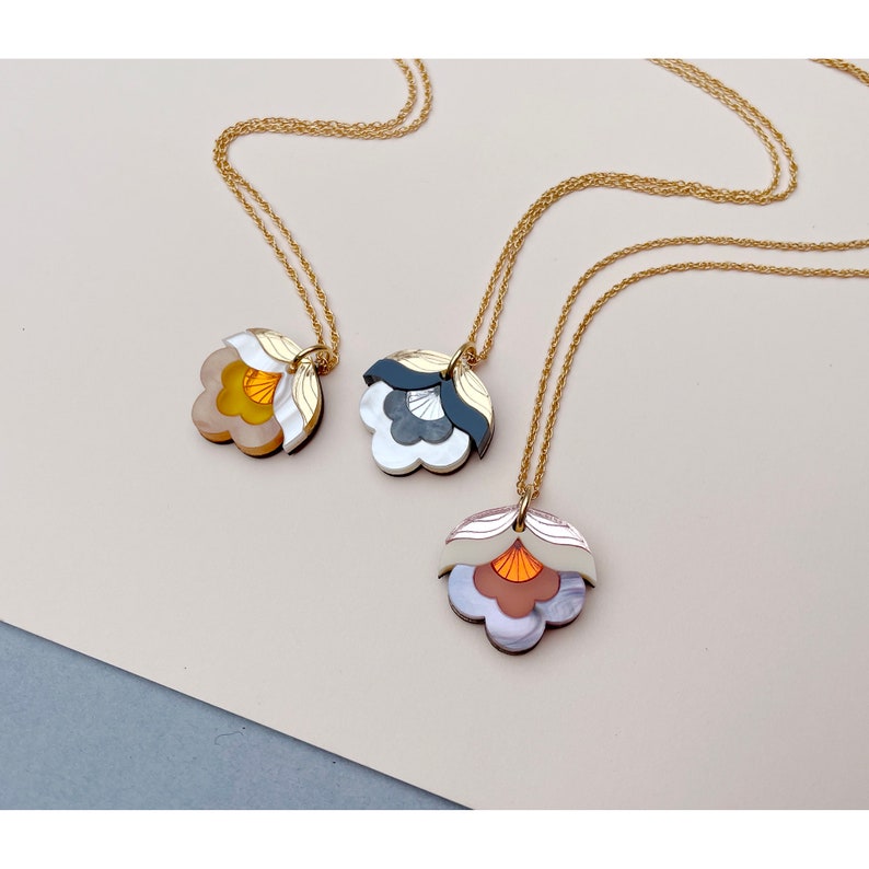 May include: Three gold chain necklaces with layered flower pendants. The pendants are made of white, black, and orange acrylic and have a geometric design.