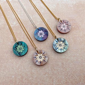Könnte beinhalten: Eine Sammlung von Halsketten mit runden Anhängern und Gold- und Silberketten. Jeder Anhänger hat ein einzigartiges Farbschema, darunter Türkis, Blau und Rosa, mit einem Sternmuster in der Mitte. Die Halsketten werden auf einer hellbraunen Oberfläche präsentiert.