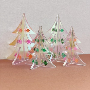 Könnte beinhalten: Vier transparente Weihnachtsbaumdekorationen aus Acryl mit bunten Sternakzenten. Die Bäume haben ein holografisches Finish, das einen Regenbogen von Farben reflektiert. Die Sterne sind in Rosa-, Grün- und Orangetönen gehalten. Die Bäume sind auf einer rosa Oberfläche angeordnet.