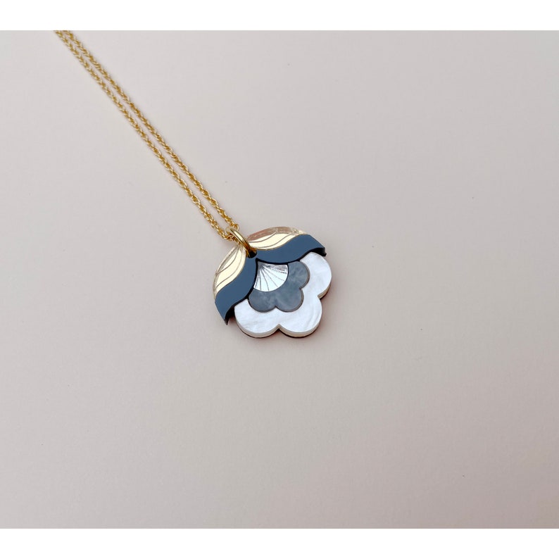 Pode incluir: Um colar de corrente dourada com um pingente em camadas que apresenta um design de flor branca, cinza e preta.