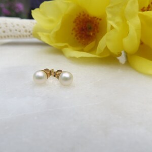 May include: A pair of pearl earrings with gold posts. The earrings are on a white surface with yellow flowers in the background.