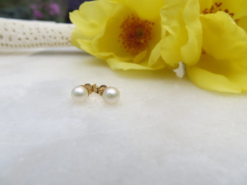 May include: A pair of pearl earrings with gold posts. The earrings are on a white surface with yellow flowers in the background.