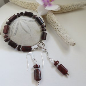Op de afbeelding: Een set rode jaspis sieraden met een armband en oorbellen. De armband is gemaakt met een zilveren toggle sluiting en de oorbellen zijn zilveren oorhaken.