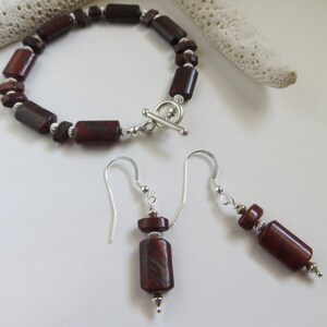 Op de afbeelding: Een set rode jaspis sieraden met een armband en oorbellen. De armband is gemaakt met rechthoekige rode jaspis kralen en een zilveren toggle sluiting. De oorbellen zijn hangend met rode jaspis kralen en zilveren haakjes.