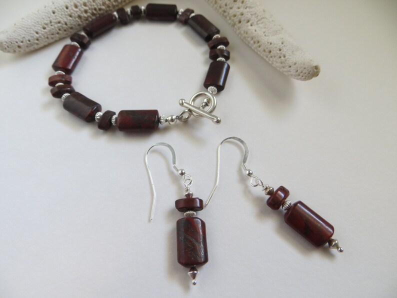 Op de afbeelding: Een set rode jaspis sieraden met een armband en oorbellen. De armband is gemaakt met rechthoekige rode jaspis kralen en een zilveren toggle sluiting. De oorbellen zijn hangend met rode jaspis kralen en zilveren haakjes.