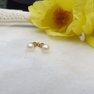 May include: A pair of pearl stud earrings with gold settings. The earrings are on a white surface with yellow flowers in the background.
