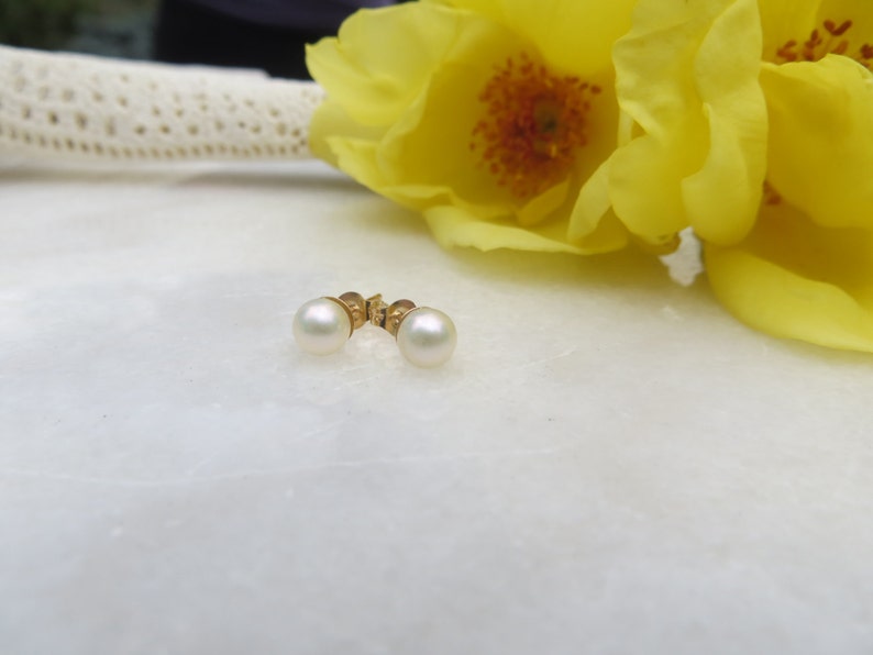 May include: A pair of pearl stud earrings with gold settings. The earrings are on a white surface with yellow flowers in the background.