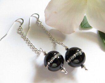 May include: A pair of dangle earrings featuring black, spherical beads with a band of clear rhinestones. The earrings have silver-colored chain and hook closures. The background is white with a white flower.