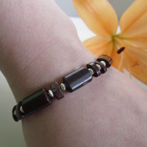 Op de afbeelding: Een bruin en zilveren kralenarmband met rechthoekige kralen. De armband wordt gedragen op de pols van een persoon.