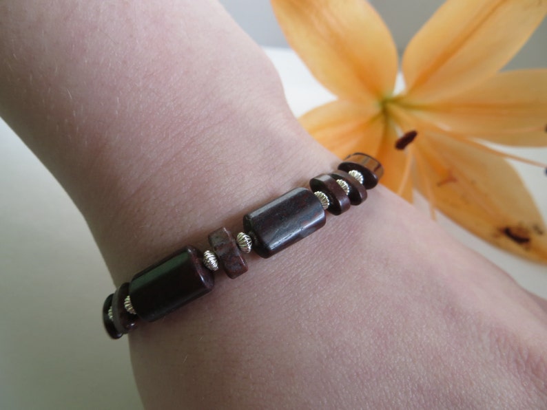 Op de afbeelding: Een bruin en zilveren kralenarmband met rechthoekige kralen. De armband wordt gedragen op de pols van een persoon.
