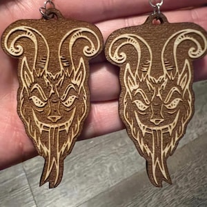 May include: A pair of wooden earrings with a detailed design of a creature with horns and a menacing expression. The earrings are brown with a light brown design.