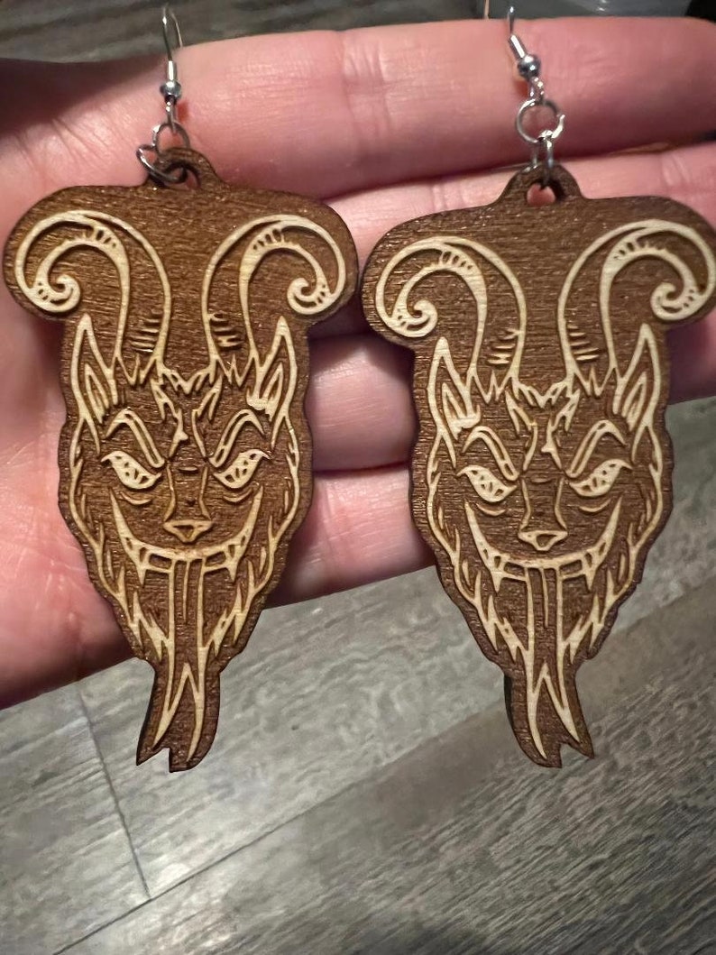 May include: A pair of wooden earrings featuring a detailed design of a creature with horns and a menacing expression. The earrings are brown with a light brown design.
