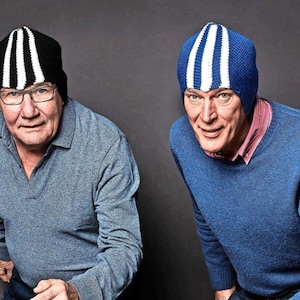 Puede incluir: Dos gorros de punto con diseños de rayas. Uno es negro con rayas blancas y el otro es azul con rayas blancas. Ambos son usados por personas que visten suéteres. El fondo es gris liso.