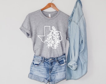May include: A gray t-shirt with a white outline of the state of Texas with a cluster of bluebonnet flowers inside. The shirt is hanging on a wooden hanger. A denim jacket is hanging next to the shirt.