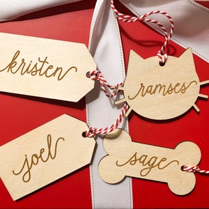 May include: Three wooden gift tags with red and white twine. The tags are shaped like a cat, a bone, and a rectangle. The cat tag says "ramses", the bone tag says "sage", and the rectangle tag says "kristen".