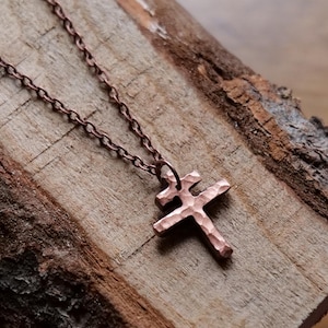 Könnte beinhalten: Eine kupferfarbene Halskette mit einem gehämmerten Kreuzanhänger. Das Kreuz mit seiner strukturierten Oberfläche hängt an einer feinen Kette. Die Halskette liegt auf einem Stück rustikalem, strukturiertem Holz und bietet einen Kontrast in Texturen und Tönen.