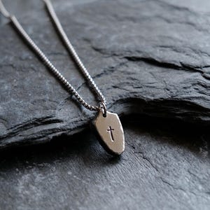 Pode incluir: Um colar prateado com um pequeno pingente em forma de escudo. O pingente apresenta um desenho de cruz estampado. O colar é exibido sobre uma superfície texturizada cinza escuro.