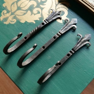 May include: Three black metal hooks with decorative fleur-de-lis finials mounted on a green surface.