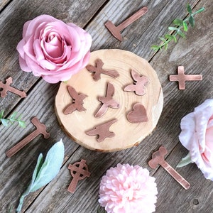 Puede incluir: Una colección de dijes de cobre en varias formas, incluyendo cruces, mariposas, un corazón y un martillo. Los dijes están dispuestos sobre una superficie de madera con flores rosas y vegetación.