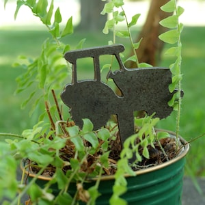 May include: A metal garden stake in the shape of a vintage car is placed in a green tin can planter filled with green plants. The background is blurred, showing a tree and grass.