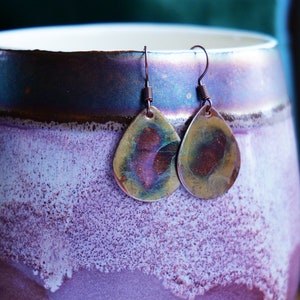 Może przedstawiać: Para kolczyków w kształcie łzy wykonanych z miedzi z młotkowanym wykończeniem. Kolczyki mają ciepły, tęczowy kolor z odcieniami złota, brązu i czerwieni.