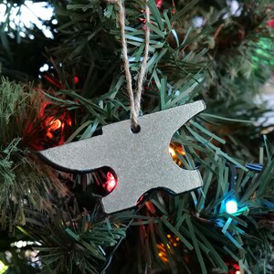 Könnte beinhalten: Ein silberfarbener, ambossförmiger Christbaumschmuck, der an einer natürlichen Schnur hängt. Der Schmuck hängt vor einem geschmückten Weihnachtsbaum mit grünen Nadeln und bunten Lichtern.