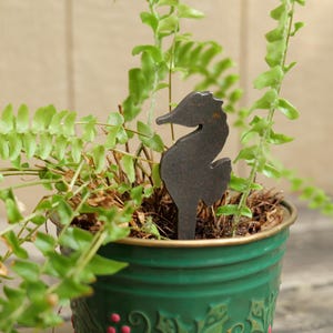 May include: A dark metal seahorse plant stake stands in a green potted plant with green foliage. The pot has a gold rim and a decorative pattern with red accents. The background is a light brown wooden surface.
