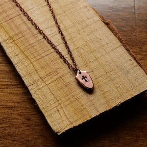 May include: A copper-colored necklace with a shield-shaped pendant featuring a cross cutout. The pendant and chain are displayed on a piece of light brown wood with visible grain. The necklace is a simple, elegant piece of jewelry.