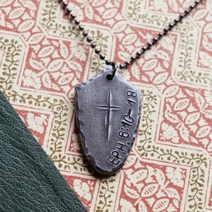 May include: A silver necklace with a pendant shaped like a shield. The pendant has a cross engraved on it and the text "EPH. 6:10-18".