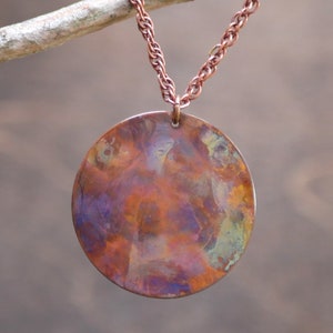 May include: A copper pendant necklace with a round, hammered disc. The disc has a colorful patina with shades of purple, green, and orange.