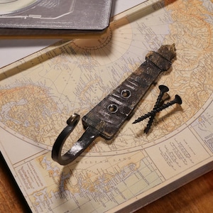 May include: A rustic metal hook with a hammered finish and a curved hook. The hook is lying on a vintage map with the text "William Land" visible. Two black screws are also visible.