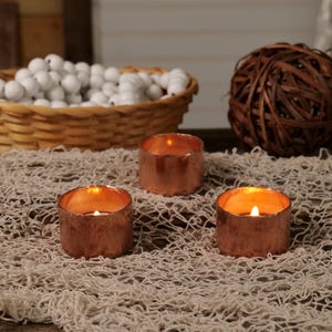 May include: Three copper candle holders with lit tea lights on a beige burlap surface.