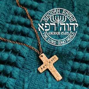 May include: A copper necklace with a hammered cross pendant. The cross has Hebrew writing on it and the words "Jehovah Rapha, Exodus 15:26, The Lord Who Heals" are written on a white circle around a menorah.