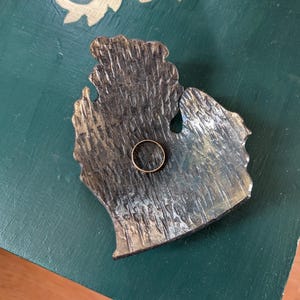 Könnte beinhalten: Ein rustikaler, dunkelbrauner, metallener Ringhalter in Form des Bundesstaates Michigan. Ein goldener Ring liegt im Halter.