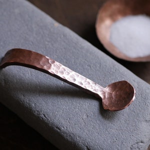 May include: A small, hammered copper spoon with a rounded bowl. The spoon is resting on a grey stone surface.