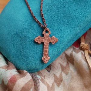 May include: A copper cross pendant necklace with a hammered texture, hanging on a turquoise blue velvet cushion.