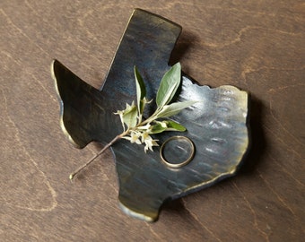 May include: A rustic metal ring dish shaped like the state of Texas. The dish is dark blue with gold accents and has a small sprig of greenery with white flowers inside.