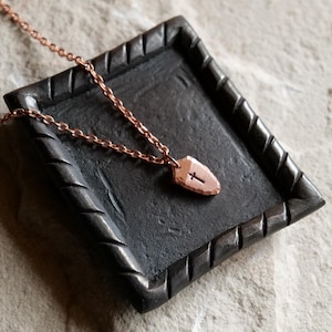 May include: A copper-colored necklace with a pendant shaped like a shield, featuring a cross. The necklace rests on a dark gray, textured tray with a decorative border. The chain is delicate and complements the pendant's design.